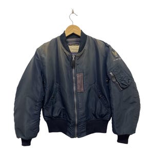 May include: A dark navy blue bomber jacket featuring a front zipper closure, ribbed cuffs and hem, and a utility pocket on the left sleeve. A circular patch with a star and wings emblem and the text US AIR FORCE is visible on the upper left shoulder.