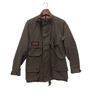 May include: An olive green field jacket featuring a high collar with a buckle strap, a front button placket, and four large flap pockets. A rectangular patch on the right chest reads PHERROW'S EXPLORATIONS. The interior collar has a plaid lining.