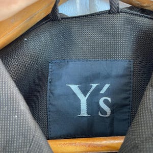 以下が含まれることがあります： ダークチャコールグレーのジャケット。襟の内側には黒い長方形のラベルがあり、白い文字でY'sとセリフ体でプリントされています。衣服は質感のある織物で作られており、襟元にはループが付いています。