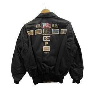 May include: A black bomber jacket featuring the text PINK HOUSE and an American flag graphic on the back. The design includes multiple rectangular patches with various emblems, the text BABY PINK HOUSE, a large letter P, and the text PINK HOUSE.