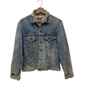 Könnte beinhalten: Eine verwaschene blaue Jeansjacke mit spitzem Kragen, langen Ärmeln mit ausgefransten Bündchen, zwei Brusttaschen mit Knopfklappen und einer Knopfleiste vorne. Der Stoff weist deutliche Abnutzungserscheinungen, Verblassungen und kleine Löcher auf.