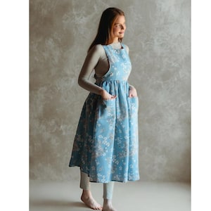May include: A light blue apron dress with a floral pattern. The dress has a scoop neckline, pockets, and a gathered skirt. The model is wearing a long-sleeved gray shirt and gray leggings.