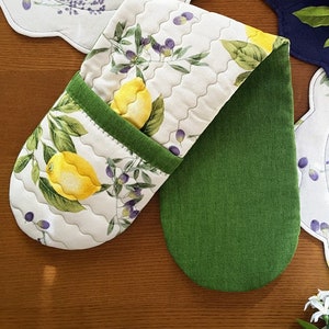 Può includere: Un guanto da forno trapuntato con sfondo bianco, con un design a limone e ramo d'ulivo. Il guanto ha un bordo verde e un retro verde unito. Il design comprende limoni gialli, foglie verdi e olive viola. Progettato per l'uso in cucina.