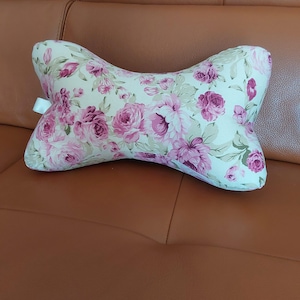 May include: A white and pink floral patterned pillow shaped like a bone. The pillow is on a brown leather sofa.