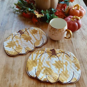 May include: Two pumpkin-shaped quilted placemats with a yellow and white leaf pattern. A white mug with gold trim and a floral design sits near two decorative pumpkins and fall foliage. The items are arranged on a wooden surface.