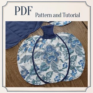 Puede incluir: Un patrón y tutorial en PDF para un artículo de tela con forma de calabaza. La calabaza presenta una base blanca con un patrón floral azul, detalles de ribete azul oscuro y un tallo azul oscuro sólido. Se ve una pieza de tela azul lisa.