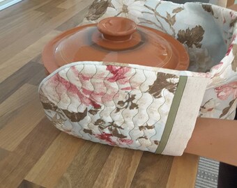 Puede incluir: Un guante de horno con estampado floral con una olla de cerámica marrón con tapa en su interior. El guante es azul claro con flores rosas y marrones. La olla está sobre una encimera de madera.