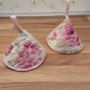Könnte beinhalten: Zwei konisch geformte, gesteppte Topflappen mit floralem Muster. Die Topflappen haben ein rosa Blumenmuster auf cremefarbenem Hintergrund. Jeder hat eine rosa Schlaufe zum Aufhängen. Sie liegen auf einer hellen Holzoberfläche.