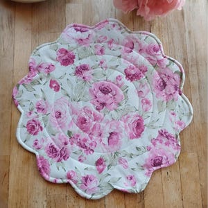 May include: A round, scalloped, quilted placemat with a floral pattern. The placemat is white with pink and green roses.