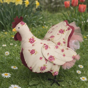 Puede incluir: Una figura de gallina de tela con cuerpo crema y alas decoradas con estampados de rosas rosas. Presenta una cresta y barbilla burdeos, un pico amarillo y patas negras. Las alas están adornadas con tul rosa. Las margaritas y otras flores están en el fondo.