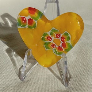 May include: A yellow heart-shaped glass ornament with a colorful geometric design. The ornament is on a clear plastic stand.