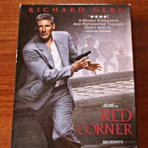 May include: A VHS video cassette cover featuring actor Richard Gere in a gray suit holding a gun. The title of the film is "Red Corner" in red letters. The tagline reads "Severity for those who resist."