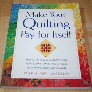 May include: A book titled "Make Your Quilting Pay for Itself" with a white cover and blue text. The book's cover features a colorful patchwork border and a diamond-shaped design. The author's name is Sylvia Ann Landman.