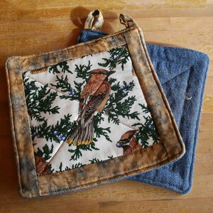 May include: Two square quilted pot holders featuring a cream-colored fabric with a pattern of brown cedar waxwing birds perched on green pine branches with blue berries. The borders are made of mottled brown fabric, and one has a blue denim backing.