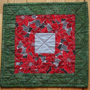 May include: A green quilted table runner with a red and white poinsettia print in the center. The runner has a white square in the center of the poinsettia print.