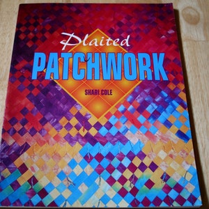 May include: A book titled "Plaited Patchwork" with a colorful geometric design cover. The cover features the title in white and blue, with the author's name, Shari Cole, below. The background is a mosaic of red, purple, orange, and blue squares.