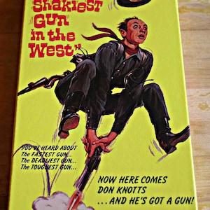 May include: A vintage VHS tape box for the movie "The Shakiest Gun in the West" starring Don Knotts. The box is yellow with a cartoon illustration of Don Knotts in a cowboy outfit, holding a gun. Text includes movie title and tagline.
