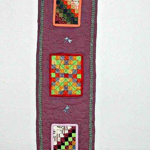 May include: A handmade, quilted wall hanging in shades of purple, with four framed patchwork squares. The squares feature colorful fabric patterns. The hanging is embellished with dragonfly motifs and a beaded trim, and is suspended from a wooden rod.