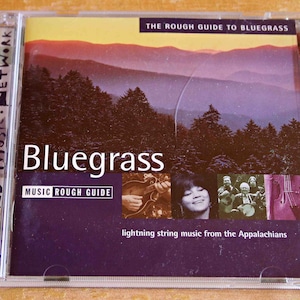 May include: A CD cover for "The Rough Guide to Bluegrass" featuring a mountain landscape and a group of musicians playing string instruments. The text "Bluegrass" is in large white letters on a purple background.