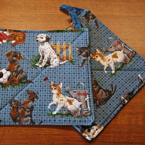 May include: Two square quilted pot holders featuring a blue plaid background with a repeating pattern of various dogs and cats. The illustrations include a dalmatian, a dachshund with a soccer ball, kittens, and garden watering cans.