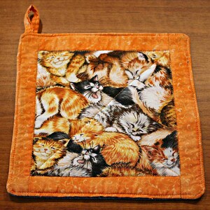 May include: A square quilted potholder featuring a central fabric panel with a dense, colorful print of multiple sleeping and playing kittens in shades of orange, black, white, and gray, framed by a solid orange border with a small fabric hanging loop.