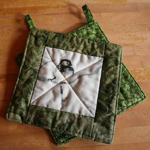 May include: A square quilted potholder featuring a central white panel with a black and white bird illustration perched on a branch, surrounded by a green fabric border with a detailed leaf pattern and a small fabric hanging loop.