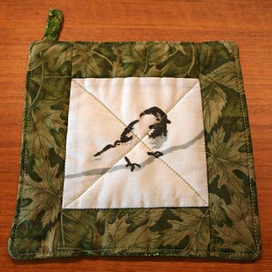 May include: A square quilted potholder featuring a central white panel with a black and white illustration of a small bird perched on a branch. The border is made of green fabric with a detailed pattern of overlapping maple leaves.