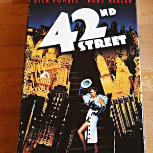 May include: A VHS tape of the musical "42nd Street" featuring Dick Powell and Ruby Keeler. The cover art depicts a woman in a white dress with black gloves and a hat, set against a backdrop of a city skyline.