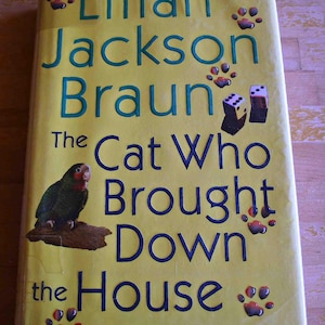 May include: A paperback book titled "The Cat Who Brought Down the House" by Lilian Jackson Braun. The cover is yellow with blue text and illustrations of a parrot, dice, and paw prints.