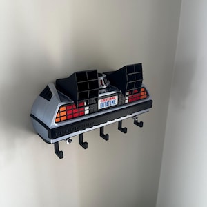 May include: A wall-mounted key holder shaped like a DeLorean car, in silver and black. The license plate reads "CALIFORNIA OUTATIME". It has four black hooks for keys. The taillights are red and orange.