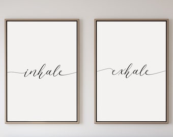 May include: Two black and white prints with the words "inhale" and "exhale" in cursive script. The prints are framed and hanging on a white wall above a bed with a light gray duvet and green pillows.