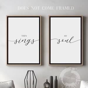 May include: Two framed prints with black text on a white background. The text on the left print reads "Then sings". The text on the right print reads "My soul". The prints are hanging on a white wall above a wooden table with two white chairs.