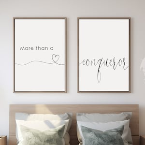 May include: Two black and white prints with inspirational quotes. The first print says "More than a" with a heart symbol. The second print says "conqueror". The prints are hanging on a white wall above a bed with white bedding and green pillows.