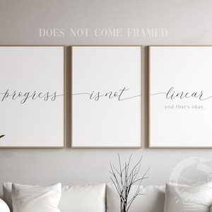 May include: Three black and white prints with the text "progress is not linear and that's okay." The prints are framed in a light wood frame and are hanging on a light gray wall.