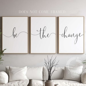 May include: Three white framed prints with black calligraphy that reads "be the change" on a white wall above a white couch with two throw pillows and a wooden coffee table.