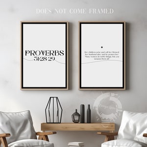 May include: Two framed prints with black text on a white background. The left print features the text "PROVERBS 31:28-29". The right print features a bible verse from Proverbs 31:28-29. The prints are displayed on a white wall above a wooden table with two chairs.