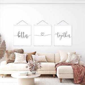 K&ouml;nnte beinhalten: Drei wei&szlig;e Leinwanddrucke mit schwarzer Kalligrafie h&auml;ngen an einer wei&szlig;en Wand. Die Drucke zeigen "better", ein Herzsymbol und "together". Die Drucke h&auml;ngen &uuml;ber einem wei&szlig;en Sofa mit braunen Kissen und einer braunen Wolldecke.