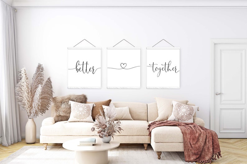K&ouml;nnte beinhalten: Drei wei&szlig;e Leinwanddrucke mit schwarzer Kalligrafie h&auml;ngen an einer wei&szlig;en Wand. Die Drucke zeigen "better", ein Herzsymbol und "together". Die Drucke h&auml;ngen &uuml;ber einem wei&szlig;en Sofa mit braunen Kissen und einer braunen Wolldecke.