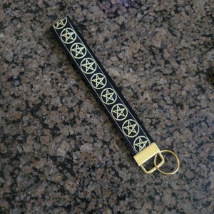 May include: A black fabric wristlet keychain featuring a repeating pattern of gold embroidered pentagrams. The strap is secured with a gold-toned metal clamp and includes a matching gold-toned circular split ring for attaching keys or accessories.