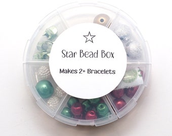 May include: A round, clear plastic storage container with six divided compartments filled with assorted beads in shades of red, green, silver, and gold. A white circular label in the center features a star icon and the text Star Bead Box Makes 2+ Bracelets.
