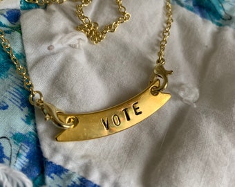 以下が含まれることがあります： 「VOTE」と大文字で書かれた湾曲したペンダントが付いた金のネックレス。ネックレスは白い布地の背景の上に置かれています。