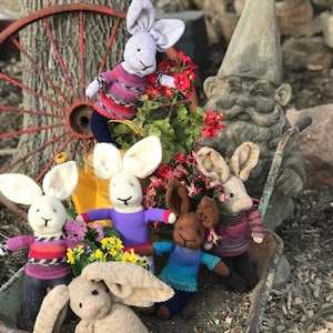 Pode incluir: Um grupo de cinco coelhos de malha em uma carriola. Os coelhos estão vestindo suéteres coloridos e estão cercados por flores. A carriola está em um caminho de terra.
