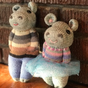 Puede incluir: Dos juguetes de ratón grises de punto, uno con un tutú azul y el otro con un suéter a rayas y pantalones azules. Los juguetes están de pie sobre una superficie de madera.