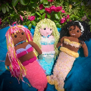 May include: Three crocheted mermaid dolls with pink, yellow, and brown hair. The dolls are wearing colorful crocheted tops and tails. The dolls are sitting on a blue fabric background.