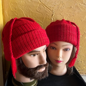 May include: Two red knitted hats with long tassels. The hats are displayed on mannequin heads. The hats are made of a ribbed knit fabric. The background is a textured gold colour.
