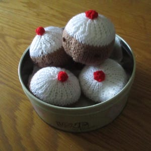 May include: A metal tin filled with four knitted cupcakes. The cupcakes have white tops, brown bottoms, and a small red pom-pom on top. The tin is light green with the words "Love to Bake" on the side.