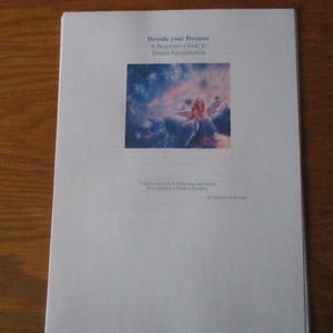 May include: A white paper with the title "Decode your Dreams" and the subtitle "A Beginner's Guide to Dream Interpretation." The image on the paper shows a woman with wings in a colorful sky. The paper is on a wooden surface.