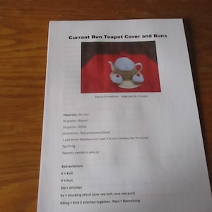 May include: A printed instruction sheet titled Currant Bun Teapot Cover and Buns. It features a photograph of a tan teapot cozy with a white top and two small bun-shaped accessories on a red surface, alongside lists of materials and abbreviations.