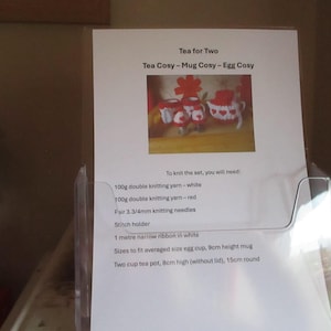 May include: A printed instruction sheet titled Tea for Two, Tea Cosy - Mug Cosy - Egg Cosy, featuring a photograph of red and white knitted covers. The text lists materials including 100g double knitting yarn, 3.3/4mm needles, and dimensions for an egg cup, 9cm mug, and 8cm high tea pot.