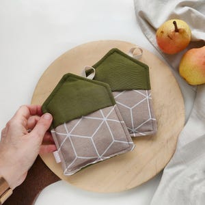 Può includere: Due presine da forno verdi e beige con un motivo geometrico. Le presine hanno la forma di una casa e hanno un anello per appenderle.